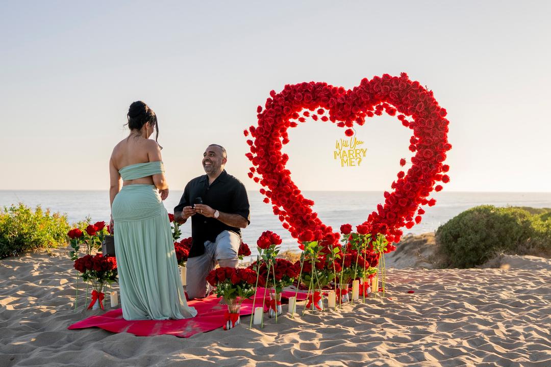He kneels with the ring beside a heart of red roses on the beach